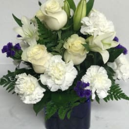 Compact arrangement of white roses, lilies, and carnations with purple accents in a dark blue cube vase