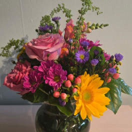 Mixed bouquet of pink roses, purple daisies, and a yellow gerbera in a glass vase
