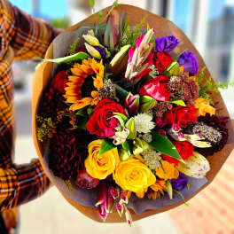 Handheld bouquet of mixed roses, sunflowers, and lilies in brown wrap