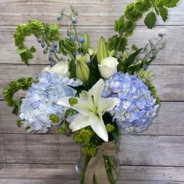 Tall arrangement of blue hydrangeas, white lilies, roses, and green bells of Ireland in a glass vase with a bow.