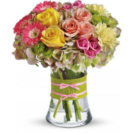 Mixed bouquet of yellow, pink, and peach flowers in a clear vase wrapped with green and pink ribbon