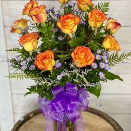 Orange-yellow roses in a glass vase with purple filler flowers and a purple ribbon