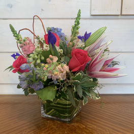 Mixed bouquet of pink roses, lilies, and blue flowers in a square glass vase