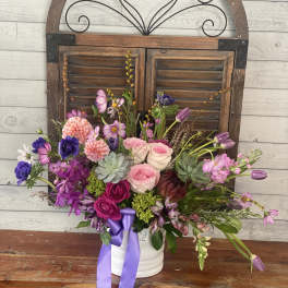 Mixed pink and purple flower arrangement in a white container with a ribbon