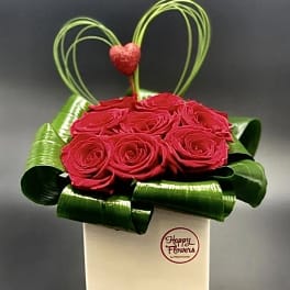 Red roses in a white box with green ribbon loops and a heart topper
