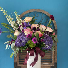 Tall floral arrangement in a white vase with purple, pink, and peach blooms