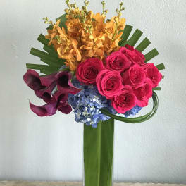Bright mixed bouquet with pink roses, orange orchids, purple calla lilies, and blue hydrangeas in a tall vase
