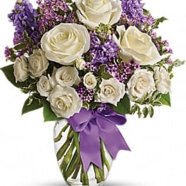 White roses and purple flowers in a clear vase with a purple ribbon