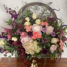 Large mixed floral arrangement in a gold vase with pink, purple, and cream blooms