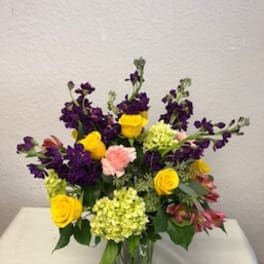 Mixed bouquet in a clear glass vase with yellow roses, purple blooms, and pink carnations