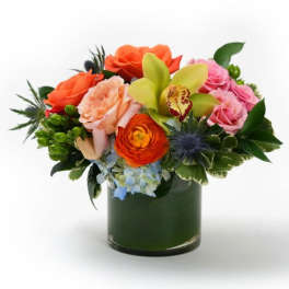 Low arrangement of orange and pink roses, green orchid, and mixed blooms in a clear glass cylinder vase.