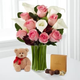 Pink roses and white calla lilies in a glass vase with a teddy bear and chocolates