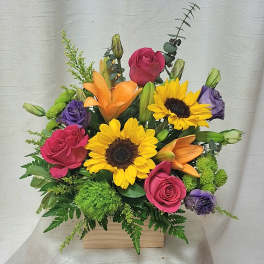 Bright mixed arrangement with sunflowers, orange lilies, pink roses, and purple blooms in a wooden box.