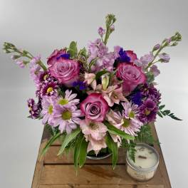 Round bouquet of pink roses, lavender mums, and alstroemeria in a glass vase beside a small candle.