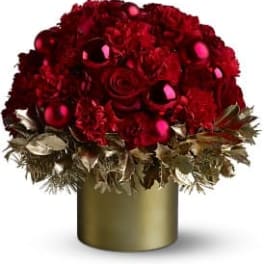 Round arrangement of red roses and carnations with red ornaments in a gold cylinder vase with gold foliage.