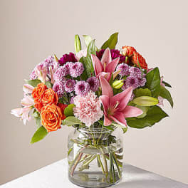 Mixed bouquet of pink lilies, orange roses, and purple mums in a glass vase