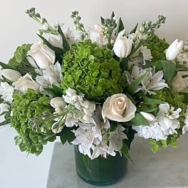Low arrangement of white roses, tulips, and green hydrangeas in a green glass cylinder vase