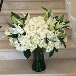 Large white arrangement of roses, lilies, and hydrangeas in a tall clear glass vase