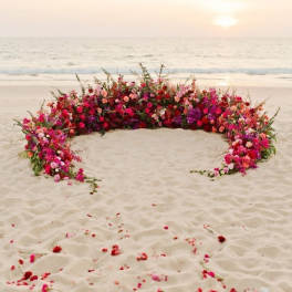 Large crescent-shaped floral arrangement on a beach at sunset