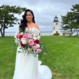 Bride holding a pink and red bouquet outdoors near a lighthouse