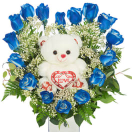 Blue roses arranged around a white teddy bear in a glass vase
