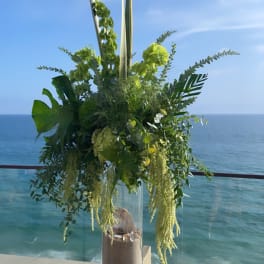 Tall green floral arrangement in a clear vase by the ocean