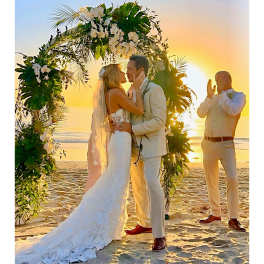 Bride and groom kissing under a floral arch on a beach at sunset