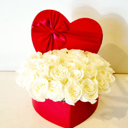 White roses arranged in a red heart-shaped box with a red ribbon bow