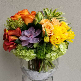 Mixed bouquet with orange roses, yellow tulips, and a purple succulent in a glass vase