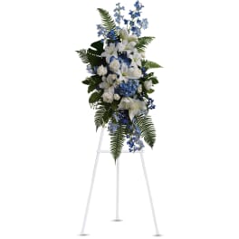 Tall standing spray of white lilies and roses with blue flowers and ferns on a white easel.