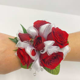Wrist corsage of red roses with white ribbon accents on a person's arm.