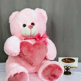 Pink teddy bear plush holding a darker pink heart pillow with a bow at its neck on a neutral backdrop