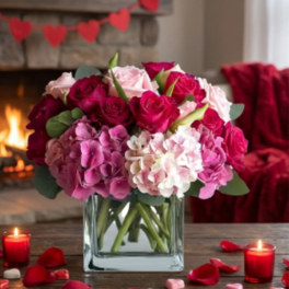 Pink and red roses with hydrangeas in a glass cube vase, surrounded by red candles and petals