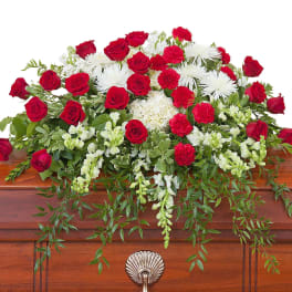 Red and white casket spray with roses, carnations, mums, and trailing greenery atop a wooden casket.