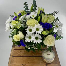 White rose, lily, and daisy bouquet with purple accents in a glass vase beside a small candle on a wooden crate.