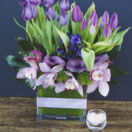 Purple tulips and pink orchids arranged in a glass vase with a small candle nearby