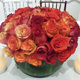 Red and orange roses arranged in a green glass bowl with two candles beside it