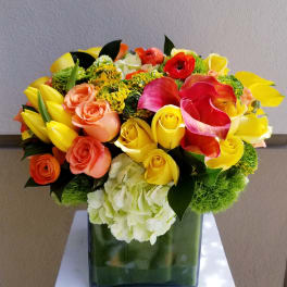 Bright bouquet of yellow roses, pink calla lilies, and orange flowers in a square vase