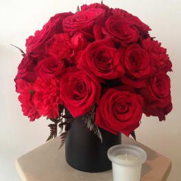 Red roses and carnations arranged in a black vase beside a white candle.