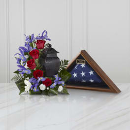 Red roses and purple irises arranged beside a folded flag in a wooden case