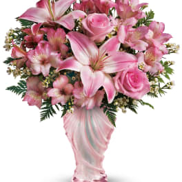 Pink lilies, roses, and alstroemeria arranged in a swirled pink glass vase