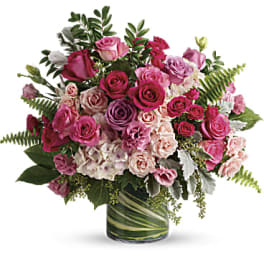Pink and lavender rose arrangement with hydrangeas in a clear glass cylinder vase wrapped with green leaves.