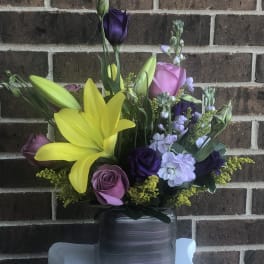 Mixed bouquet with yellow lilies, purple roses, and lavender blooms in a glass vase