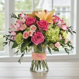 Mixed bouquet of pink roses, lilies, and green hydrangea in a clear glass vase with a pink-tied band.