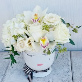 White floral arrangement in a face-shaped vase with a striped ribbon