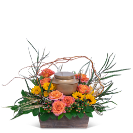 Orange roses and yellow flowers arranged around a bronze urn in a wooden container