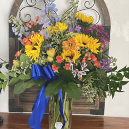 Mixed bouquet with sunflowers, purple blooms, and a blue ribbon in a glass vase