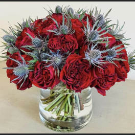 Red roses arranged in a clear glass vase with blue thistle accents.