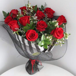 Hand-tied bouquet of red roses with white filler flowers wrapped in black mesh, with rose petals on a table