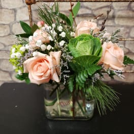 Short arrangement of peach roses, white filler blooms, and green ornamental cabbage in a glass cube vase